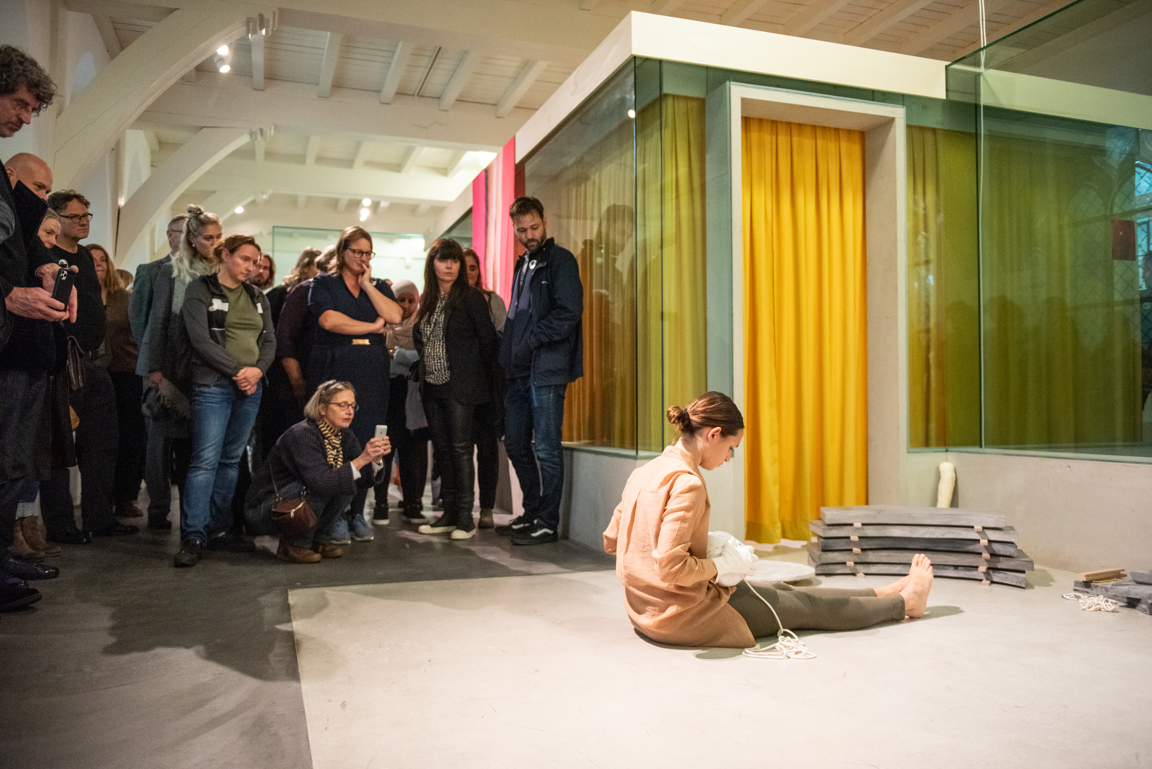bezoekers bekijken de performance in de tentoonstelling, de performer zit op de grond met een gipsen hoofd op schoot